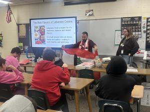 A student in a classroom presents Lebanese cuisine, holding a Lebanese flag while speaking.