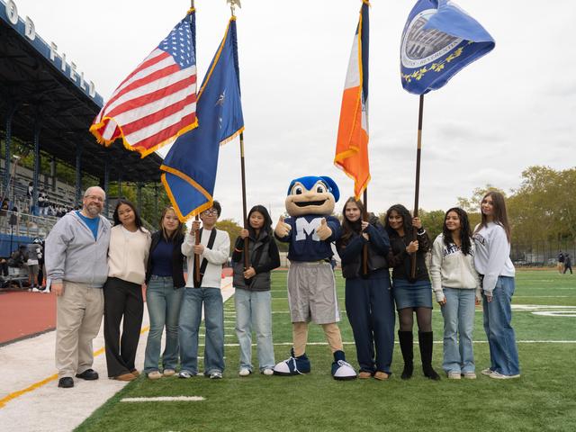 Group of people holding flags with a mascot on a sports field.