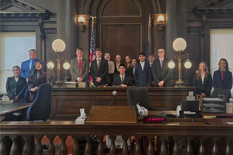 The mock trial team gathers in the courthouse sitting and standing at the judge's bench
