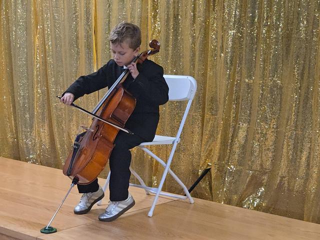 A boy in formal wear plays the cello while seated on a chair.