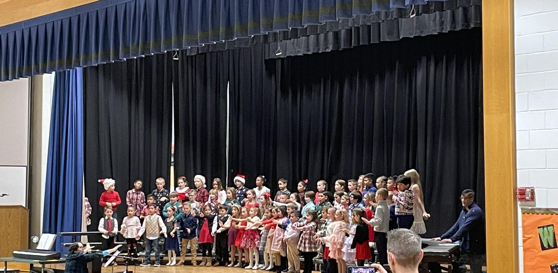 A large group of children in festive attire performing on stage with a musician at a piano.