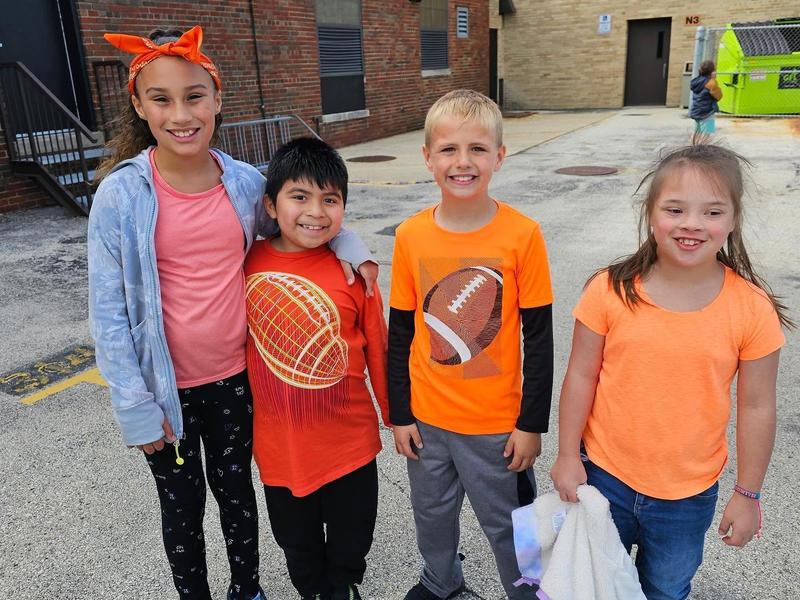 Four children smiling and posing together, wearing bright orange and casual clothes.