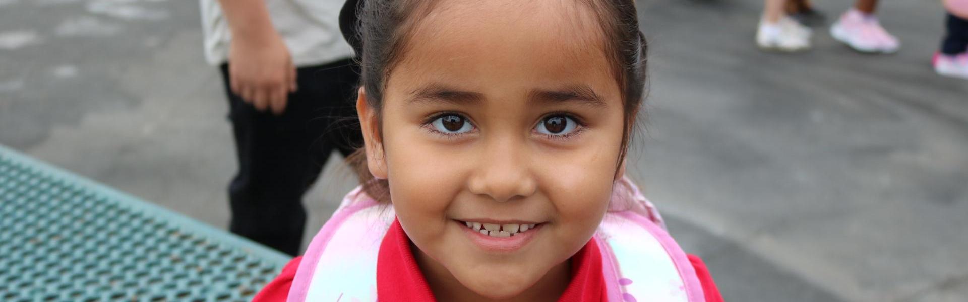Smiling young girl with a pink backpack and red shirt.