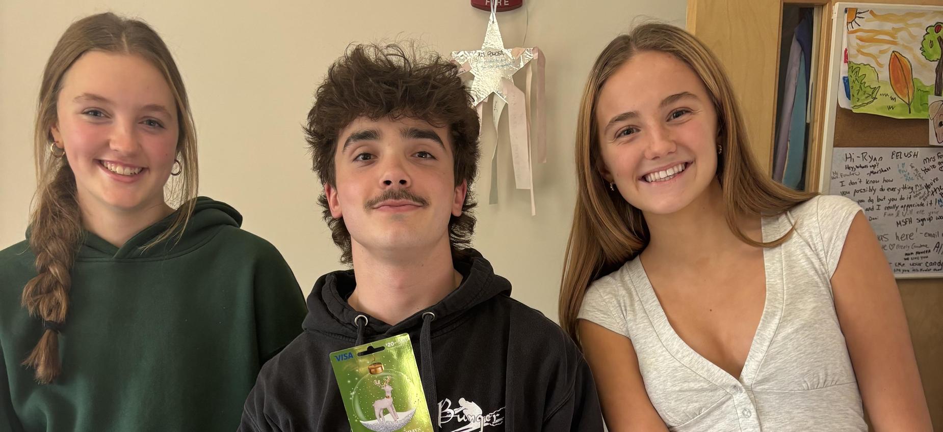 Three teenagers stand together, one holding a green flyer, wearing casual clothing.