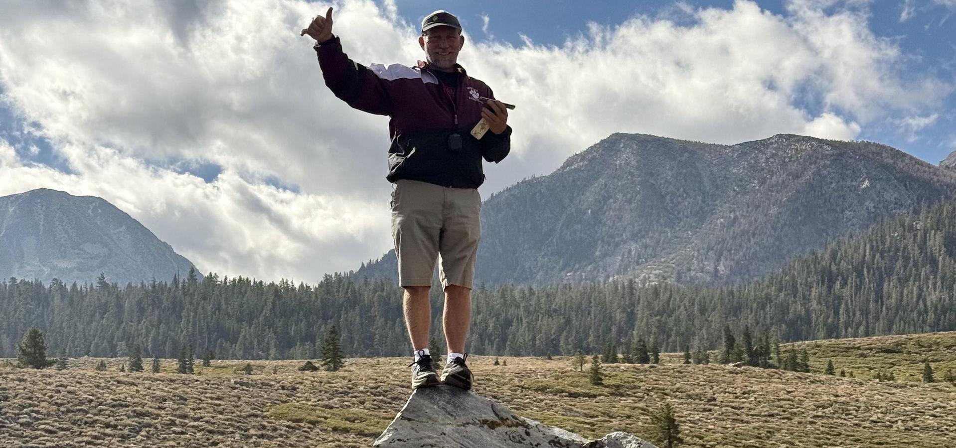 A person standing on a large rock in a mountain landscape, holding a camera.