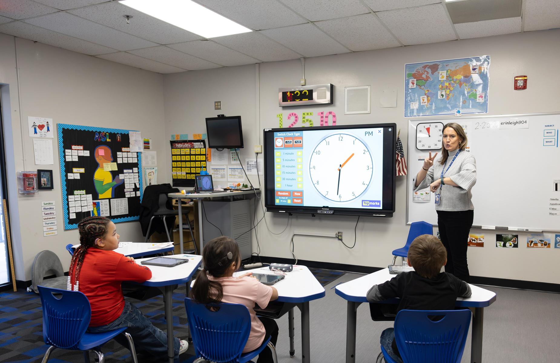 A teacher instructs students in a colorful classroom with a digital clock and map.