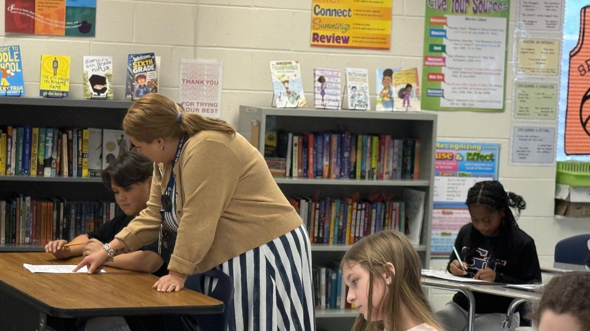 A teacher assists students in a classroom filled with books and colorful posters.