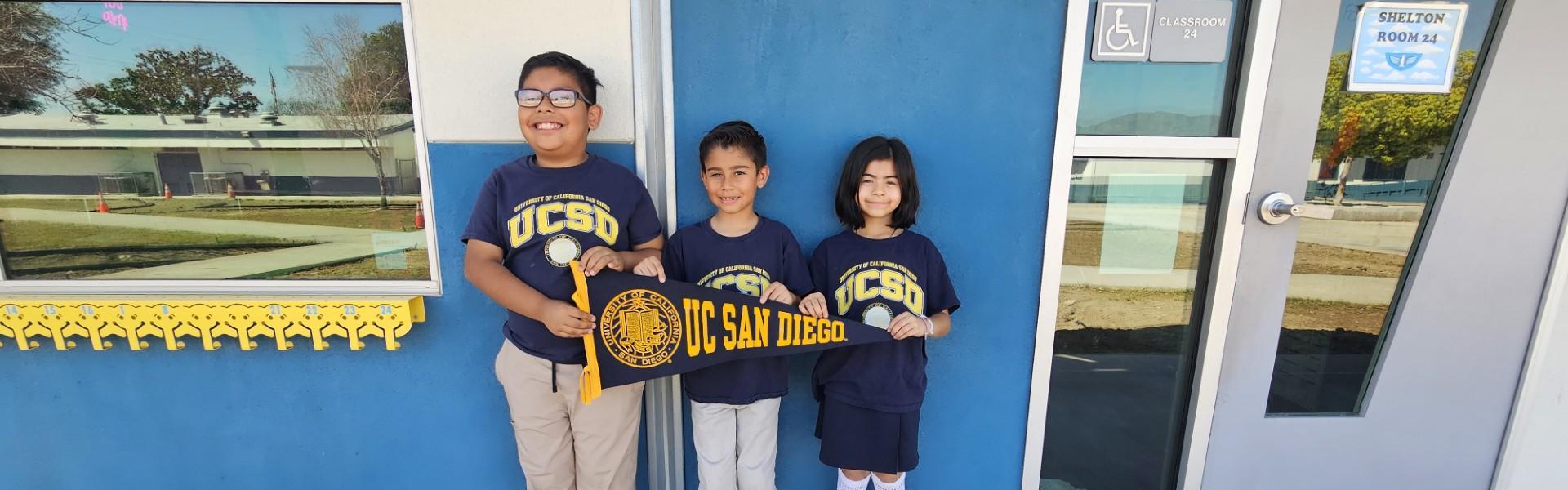 Three children pose with a UC San Diego banner in front of a blue wall.