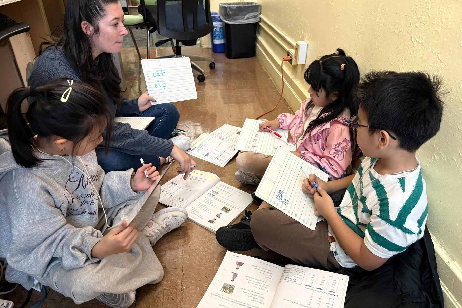 Teacher and students engaged in writing activities, using materials on the floor.