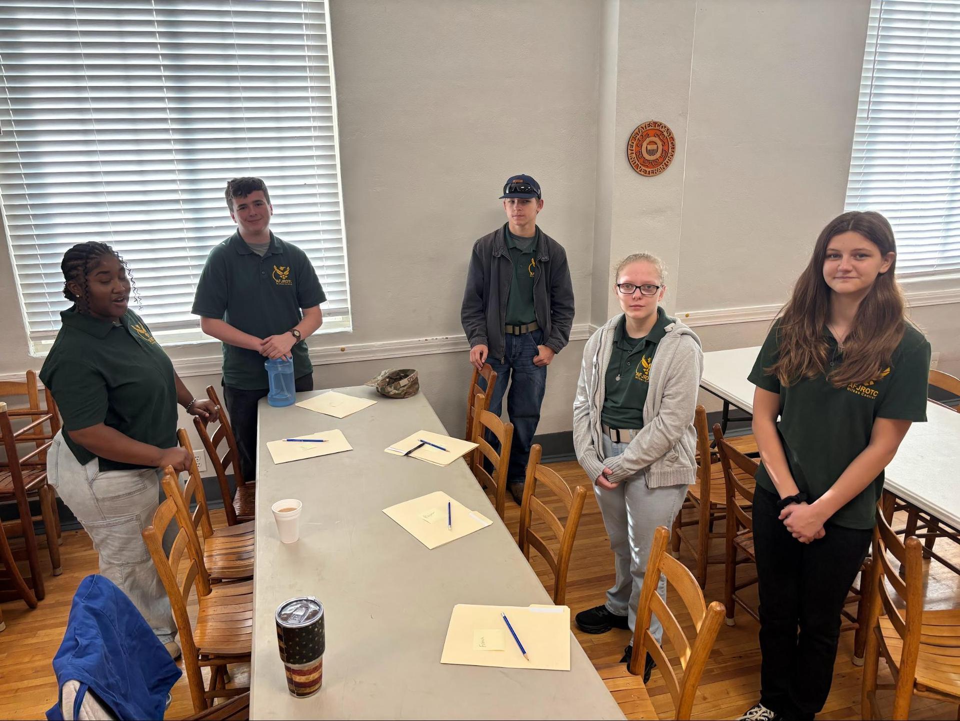 Five people standing around a table in a classroom setting with papers and drinks.