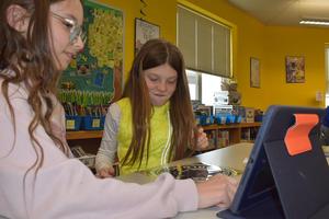 Two girls looking at a tablet and engaging with items on a table in a classroom.