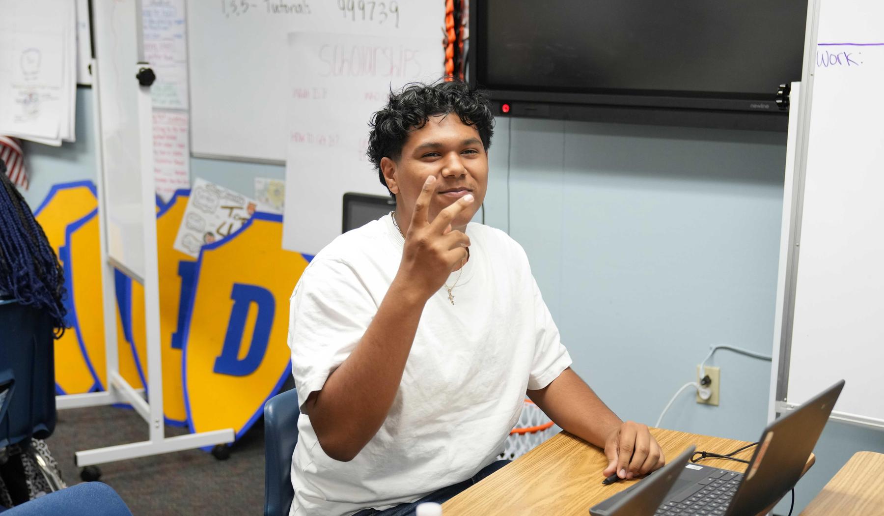 A student sitting at a desk, making a peace sign with one hand.