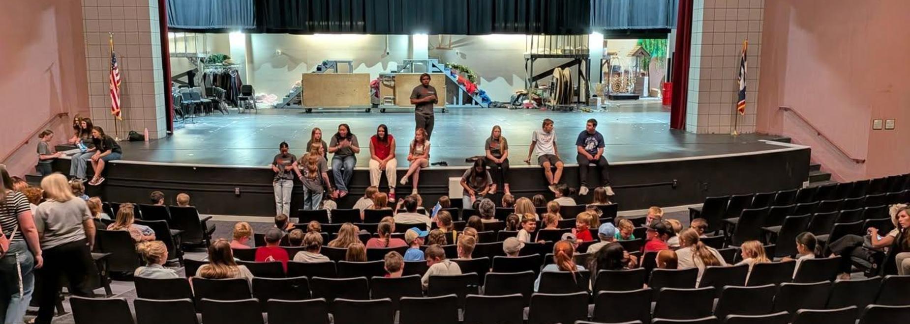 Students gather on stage in a theater with a backdrop and props visible.
