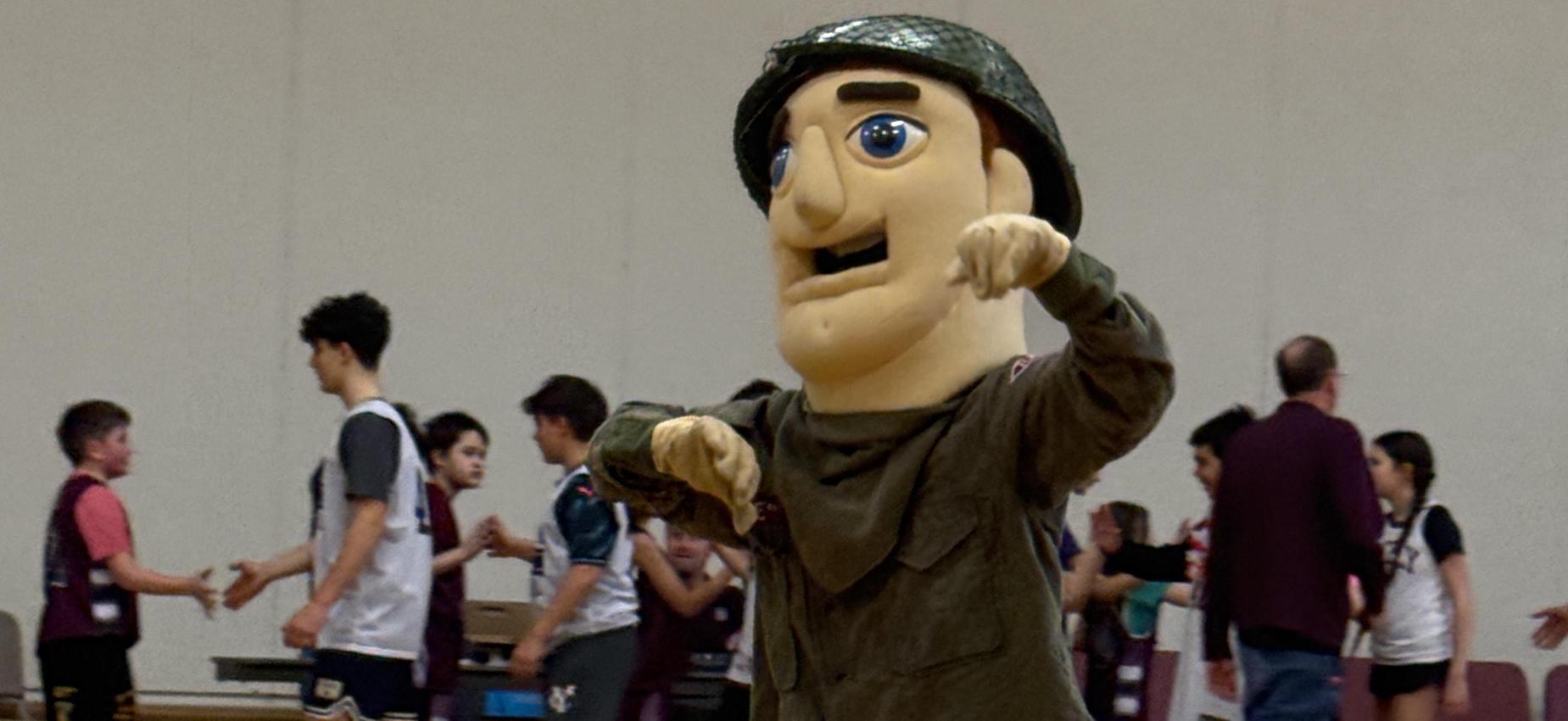 A mascot in a military uniform poses on a gym floor while players engage in a game.
