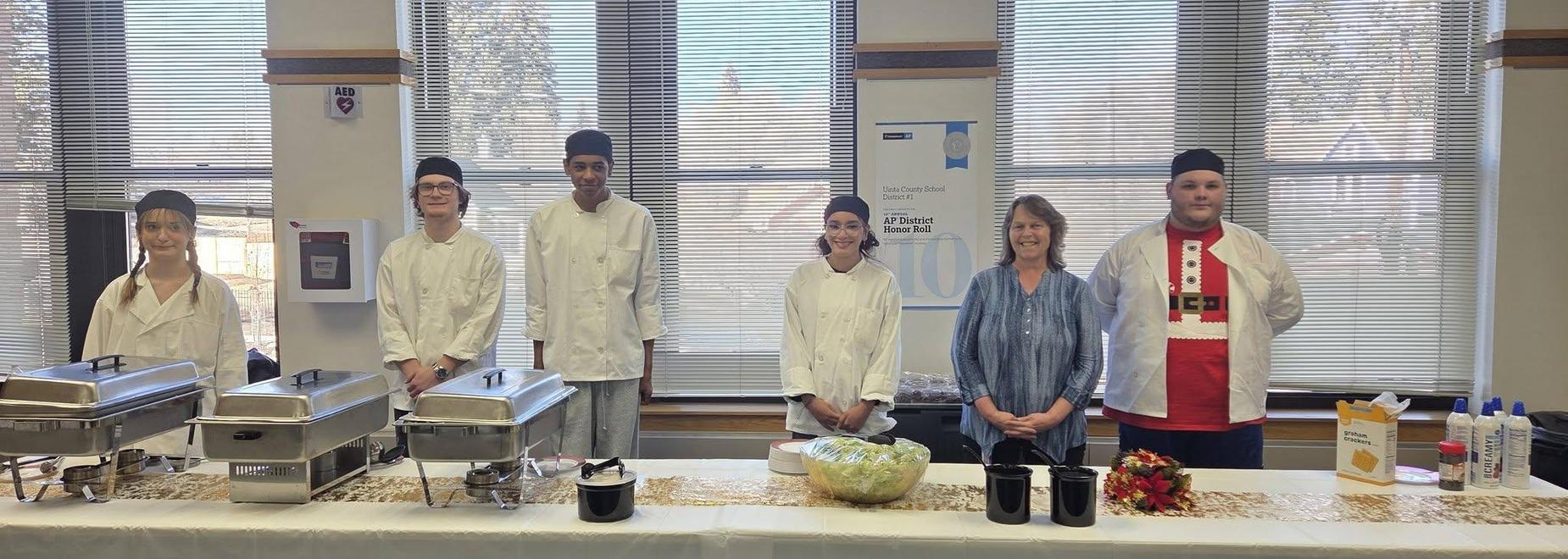 Culinary students in chef uniforms pose behind a serving table with food dishes.