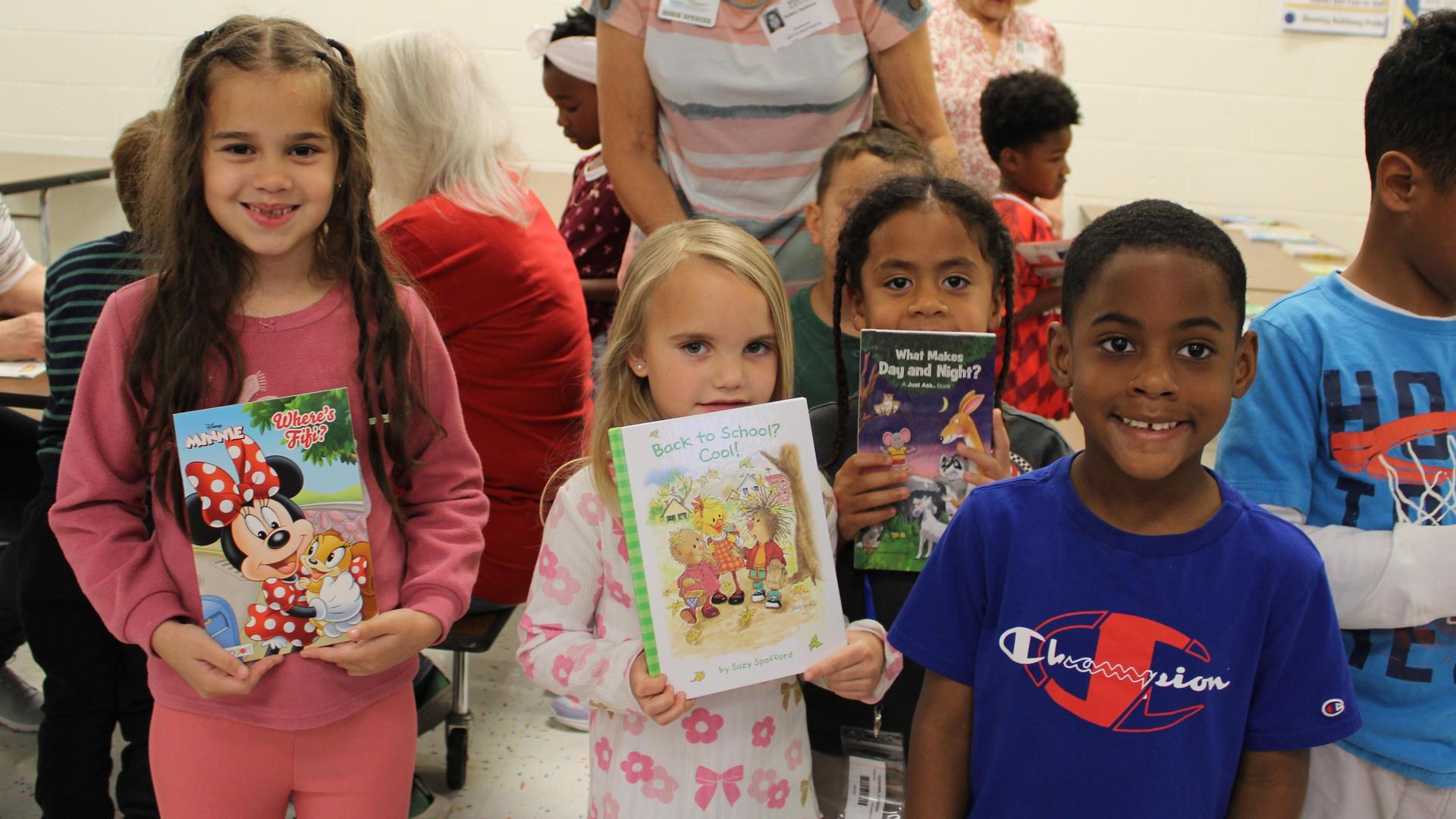 Four children holding books and smiling inside a classroom.