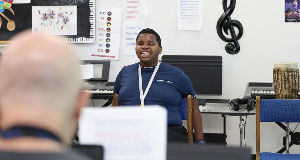 A smiling young boy sits in front of a music classroom, engaging with others.