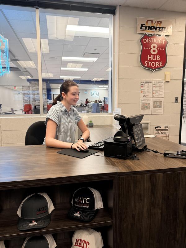 A young woman sits at a counter with a computer in a store setting.