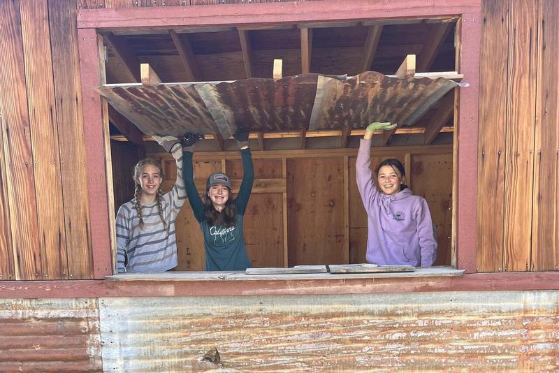Three middle school students wearing gardening gloves smile from inside a shed