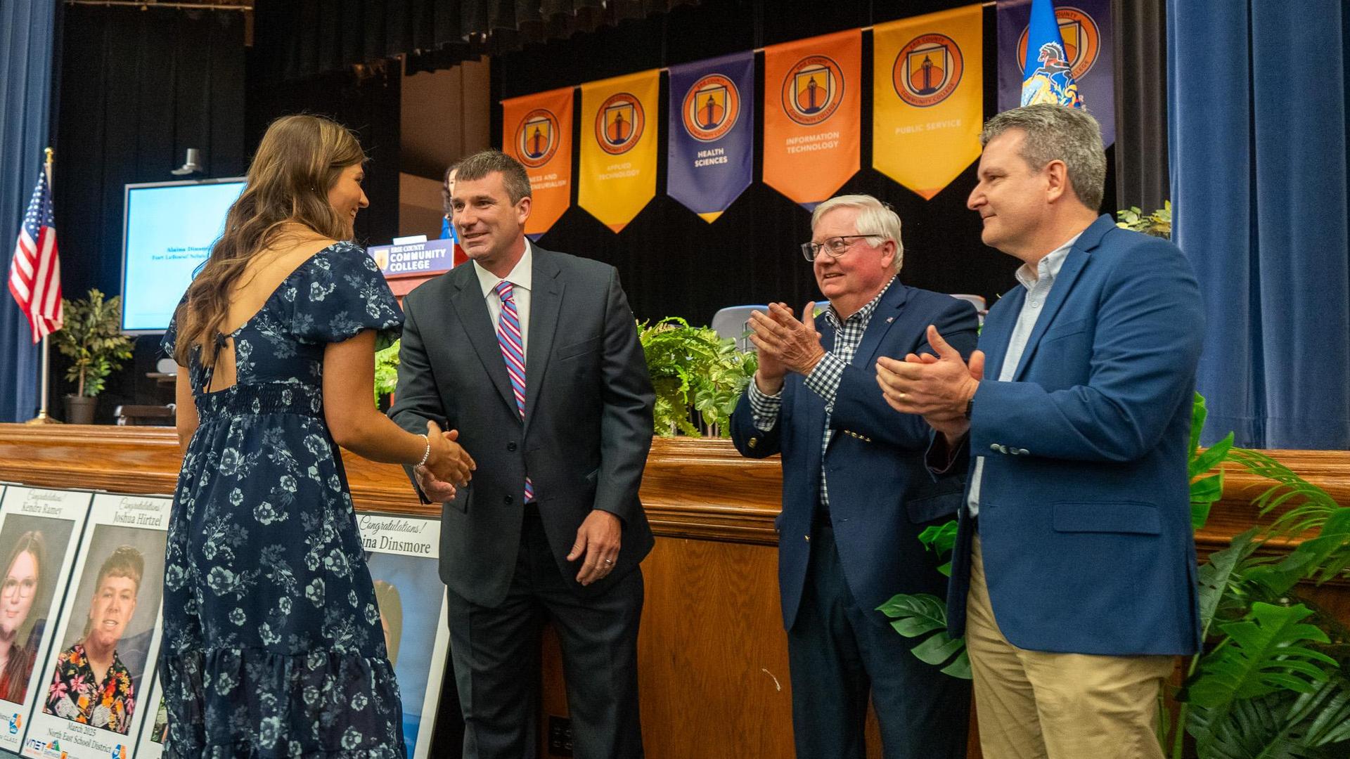 Executive Director Brad Whitman congratulates a student in a blue dress with a handshake during the Class Act Scholarship Ceremony. Two men representing organizations sponsoring the scholarship stand nearby applauding as the recognition takes place on a stage decorated with banners and plants.