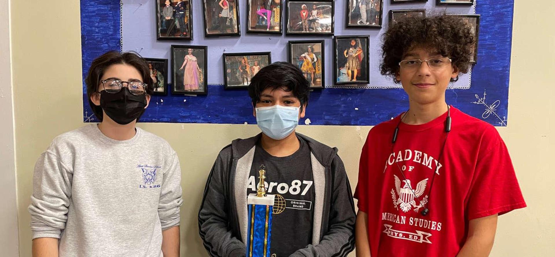 Three students holding a trophy in front of a wall of framed photos.