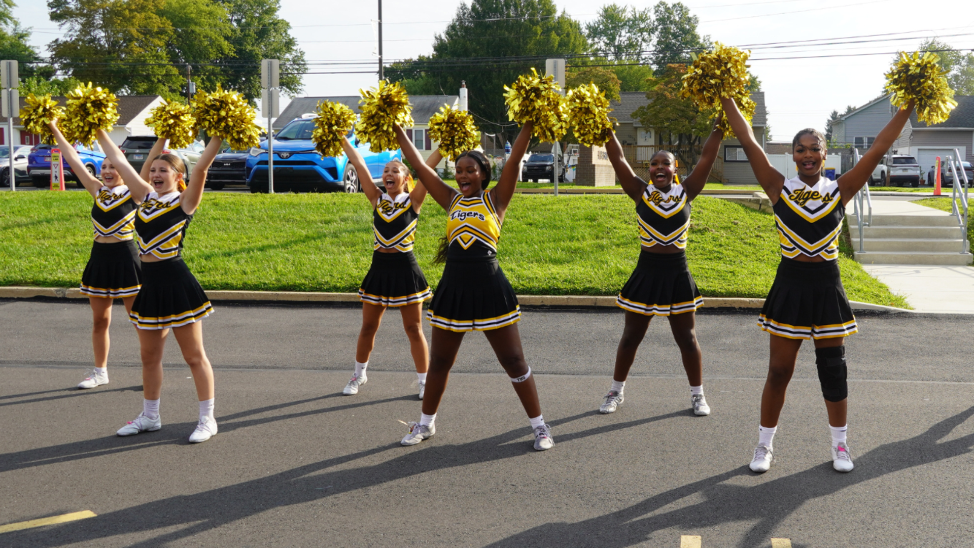 Six cheerleaders in black and gold "Tigers" uniforms stand in a line on an asphalt lot, smiling and holding gold pom-poms high in the air.