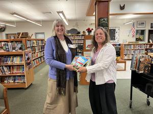 Two women exchange a book in a library filled with book shelves.