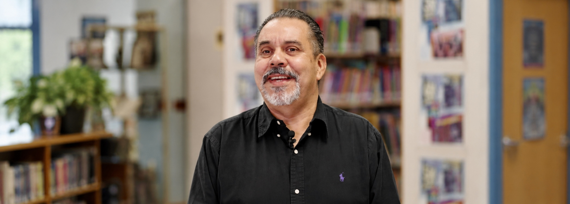 A smiling man in a black shirt standing in a library with bookshelves.