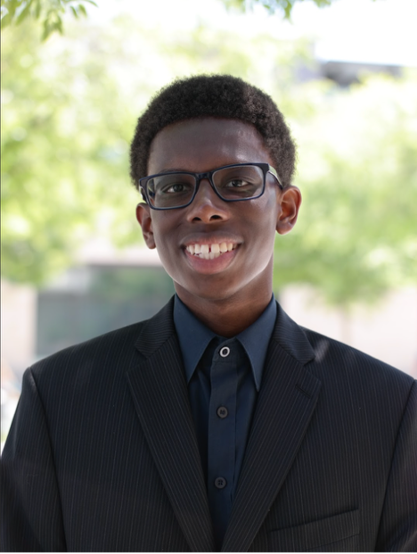 Teen boy with glasses wearing a suit, smiling outdoors.