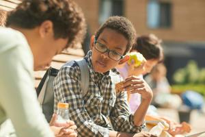 Two students sit outside on building steps eating their lunches.