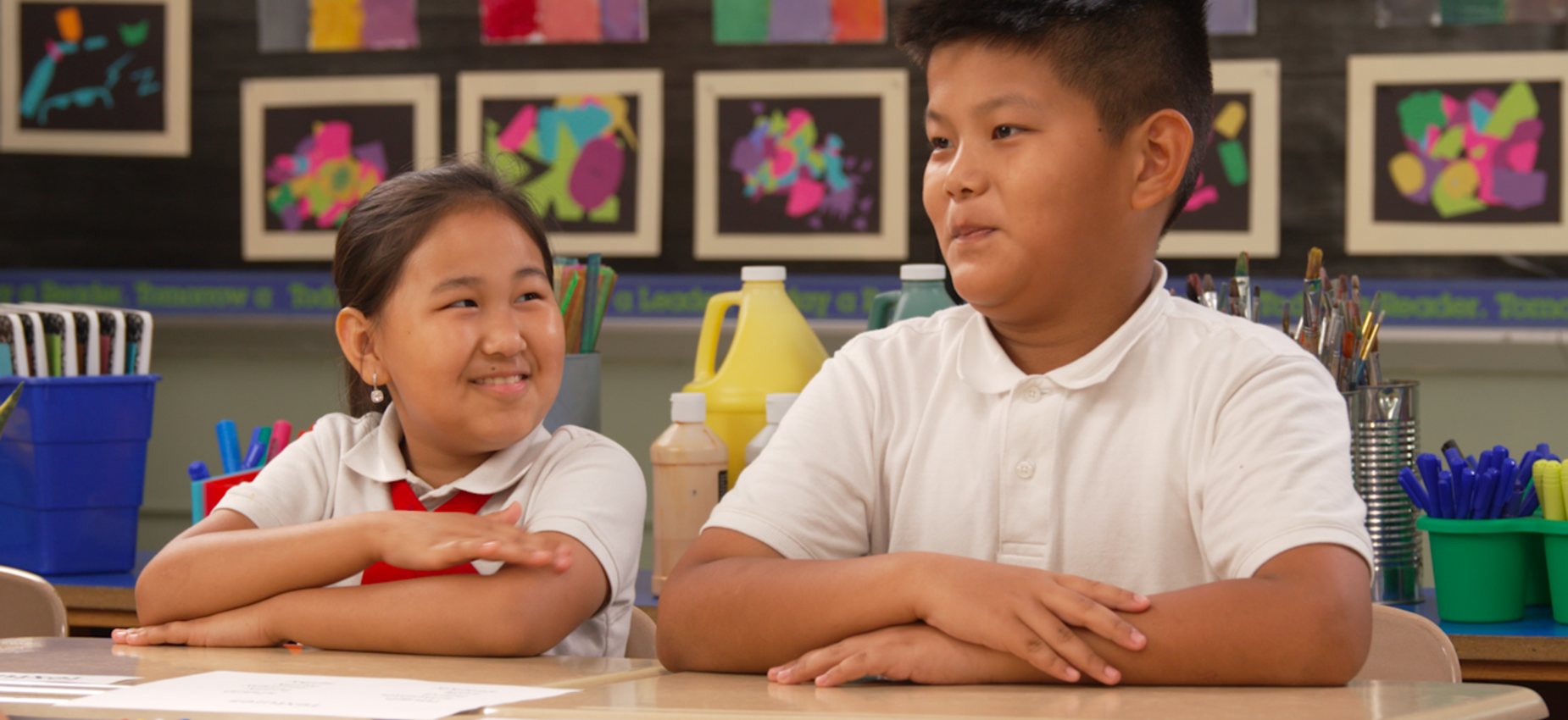 Two children sitting at a desk in a classroom, smiling and looking at each other.