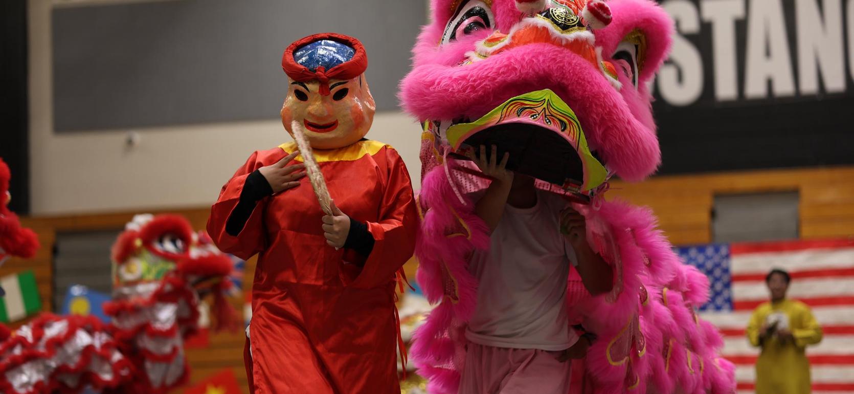 Two performers in colorful outfits participate in a lion dance event inside a gymnasium.