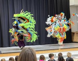 Native American dancers perform in traditional dress on a school stage.