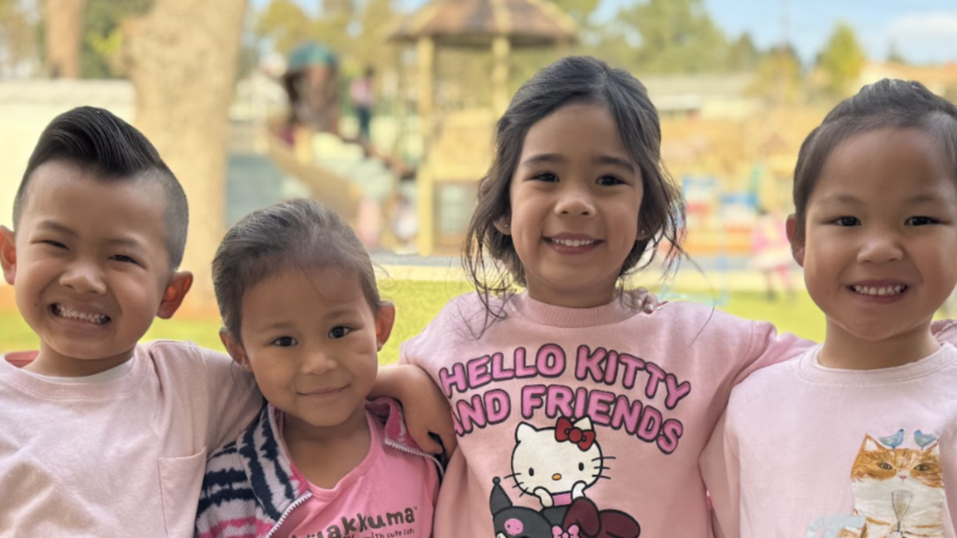 Four children smiling together outdoors in pink shirts.