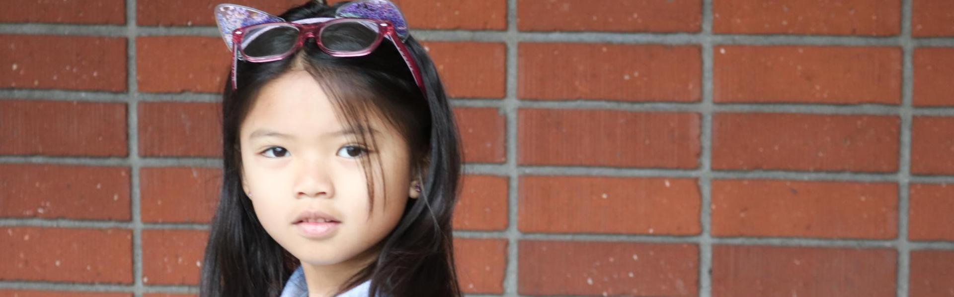 Girl with long hair, wearing sunglasses, posed against a brick wall.