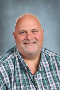 Smiling man with short hair and beard in a checked shirt against a blue-gray backdrop.