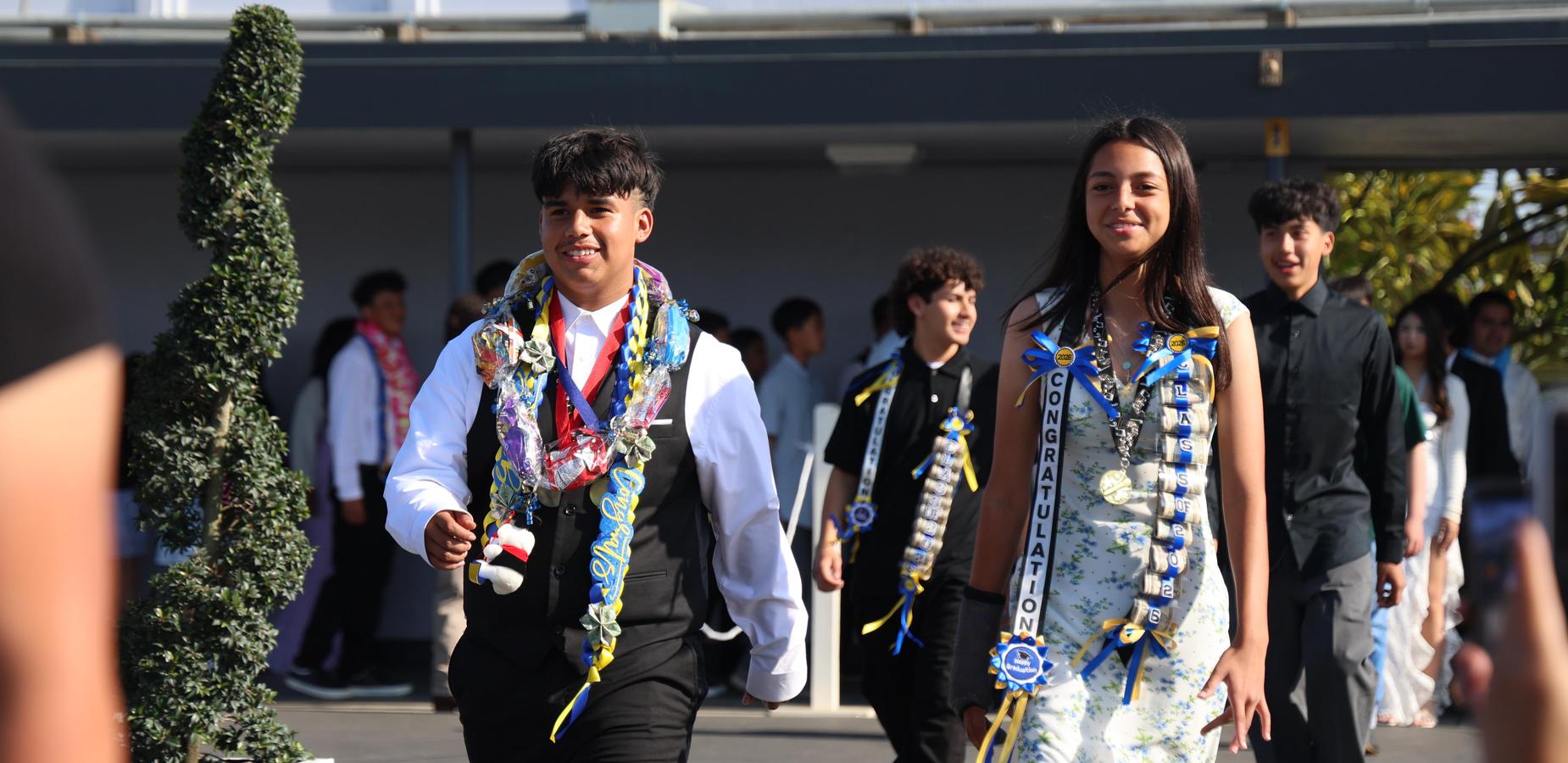 Graduates in formal attire walking with leis at a ceremony.
