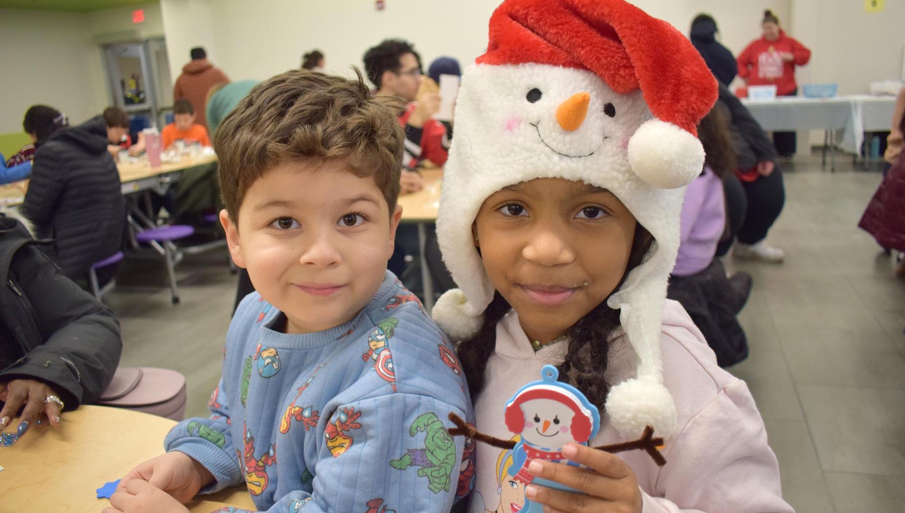 A boy and girl, both wearing winter hats, show off their crafts in a busy festive setting.