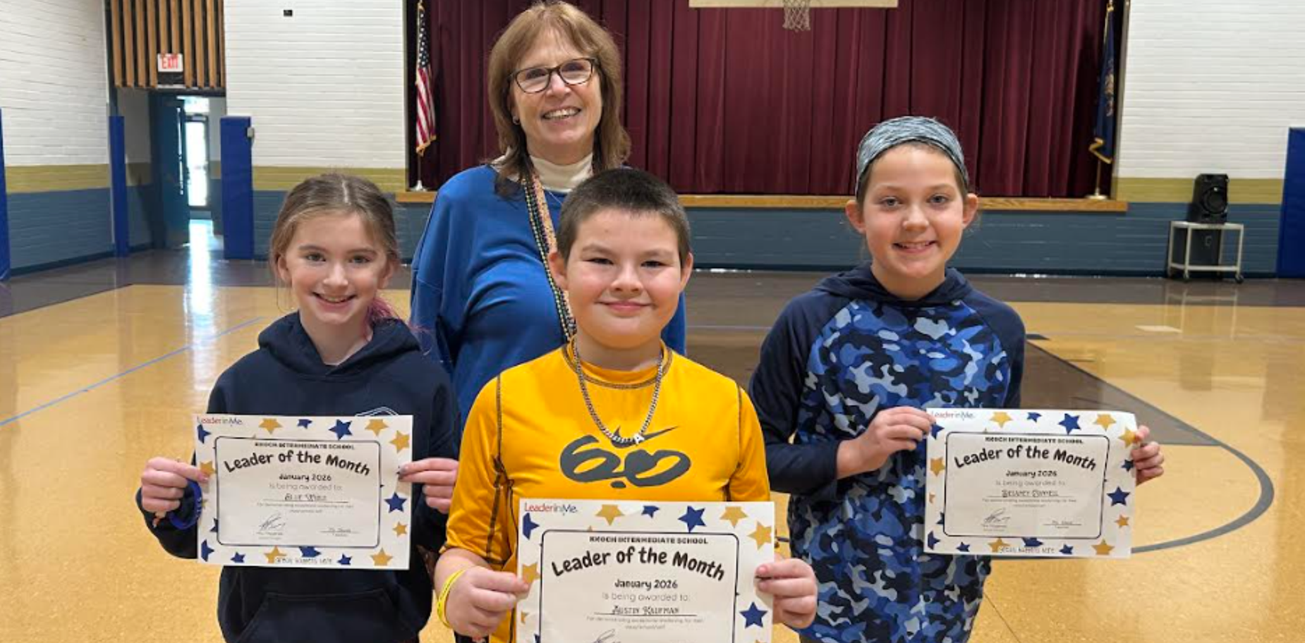 Three students holding certificates with a teacher in a gymnasium.
