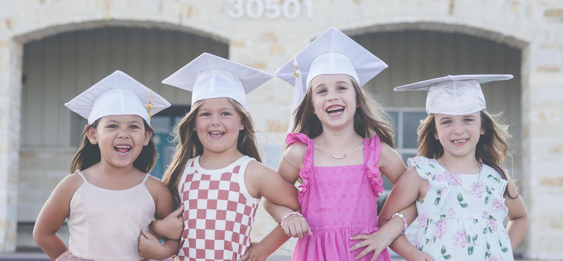 Four girls in graduation caps smiling and holding hands in front of a building.