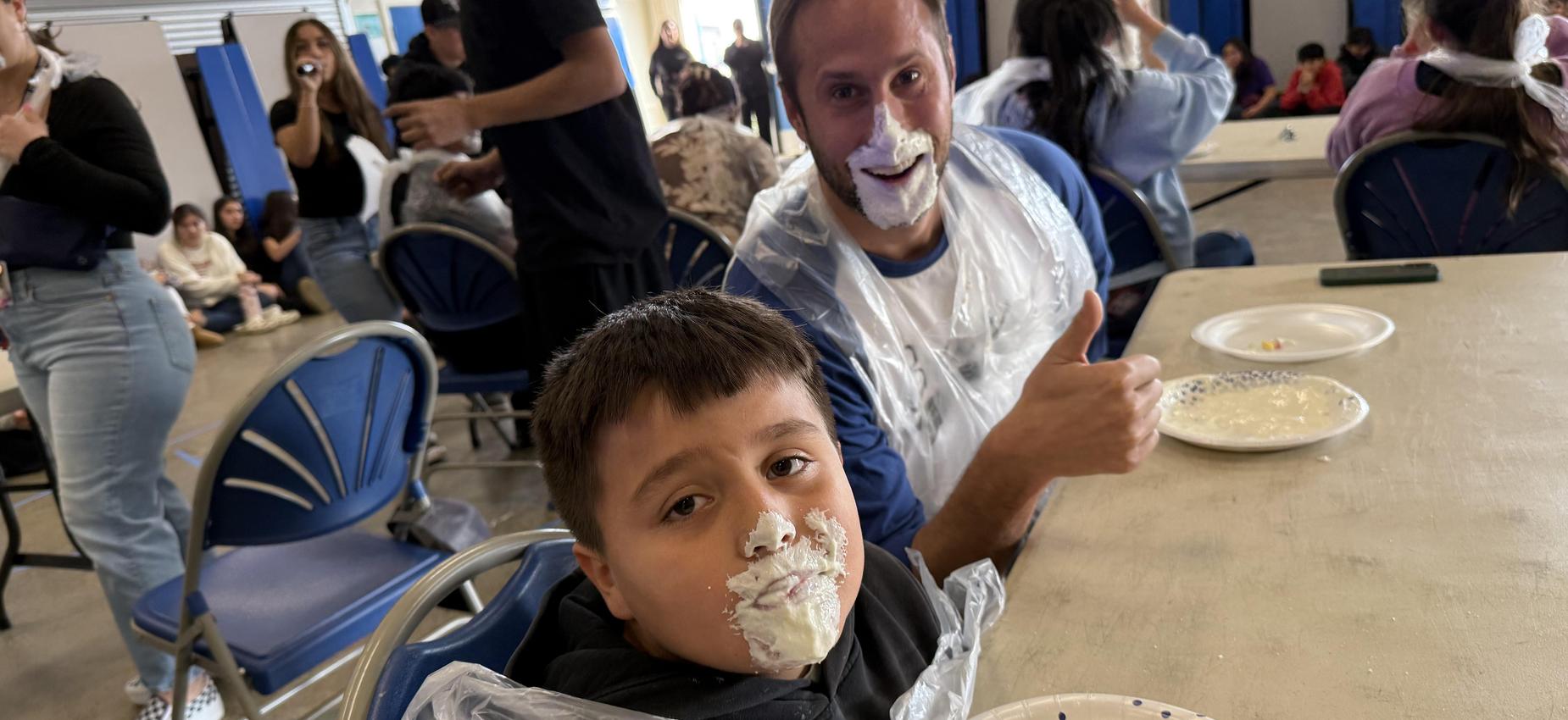 Child and adult laughing with whipped cream on their faces at a festive gathering.