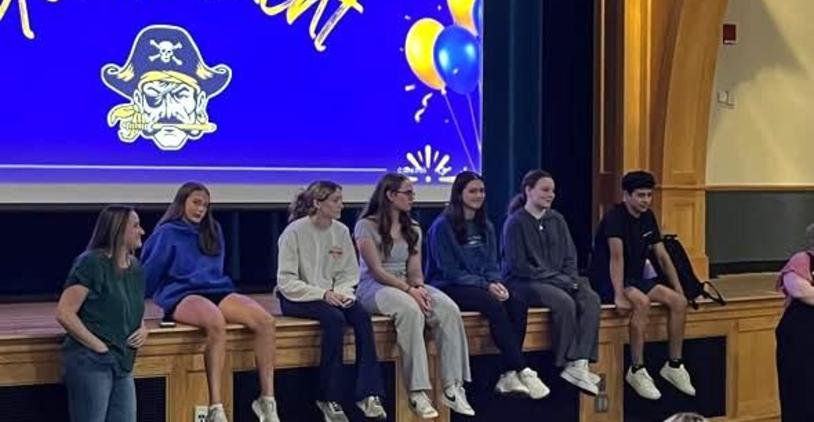 Students sitting on a stage during an event with a decorated backdrop.