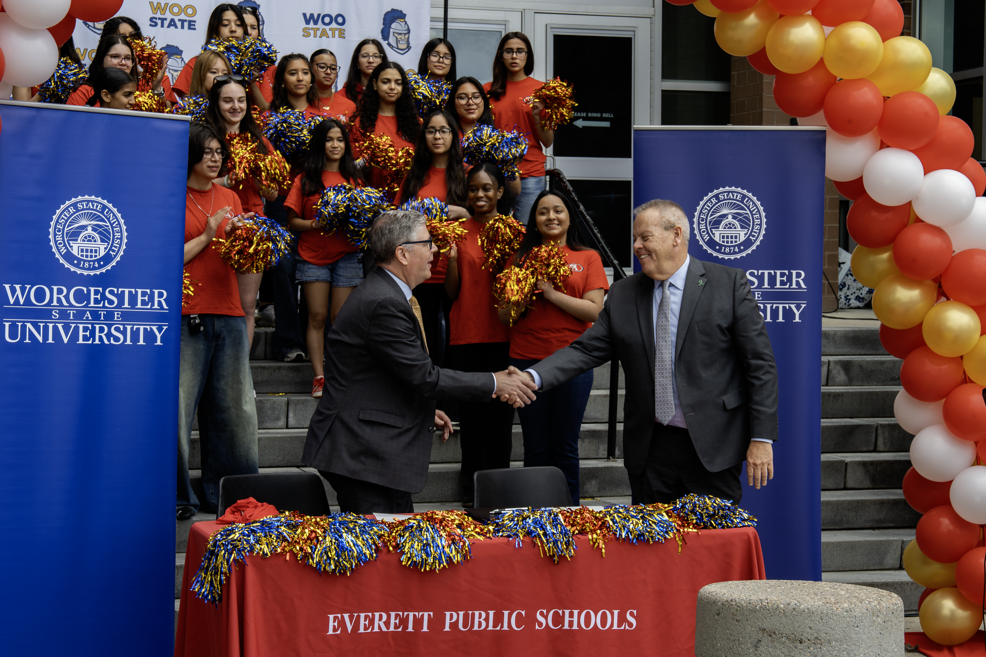Two men shake hands at a ceremony, with students holding pom-poms in the background.