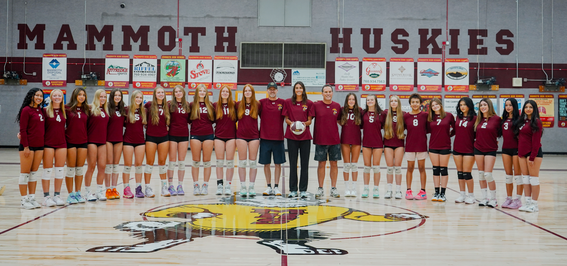 Varsity volleyball team posed together on a gym floor with team banners behind them.