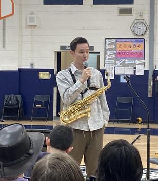 Man wearing a patterned shirt speaks into a microphone while holding a saxophone.