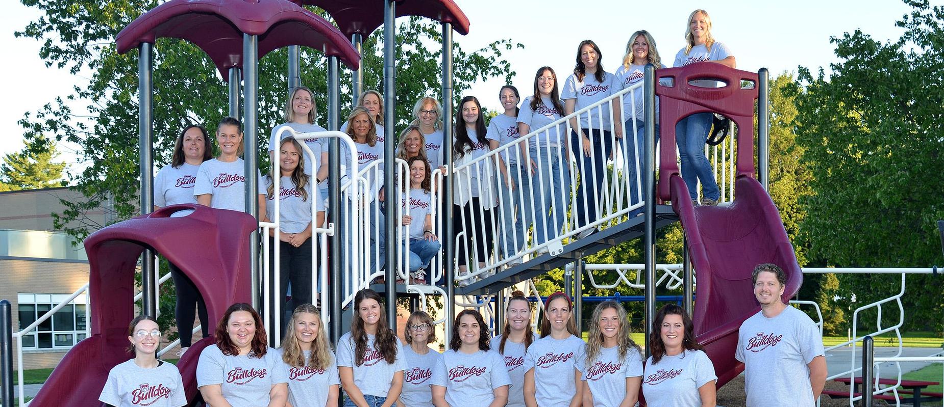 staff pose in grey Bulldog Tshirts in front of a slide on the playground as well as on the top walkway