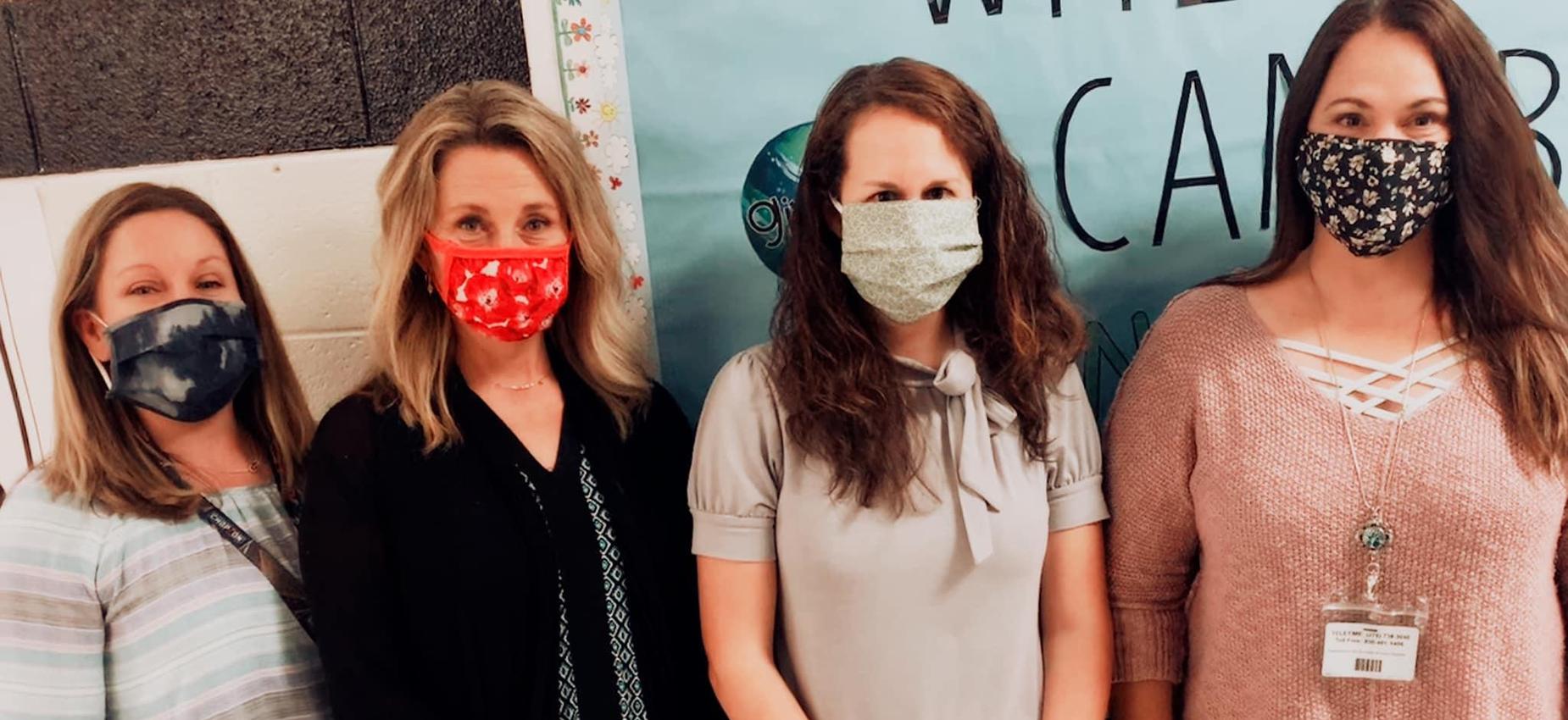 Four teachers wear masks in front of a bulletin board.