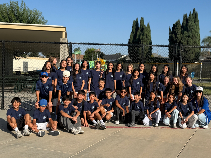 Group of students in matching navy t-shirts posing outdoors by a school fence.