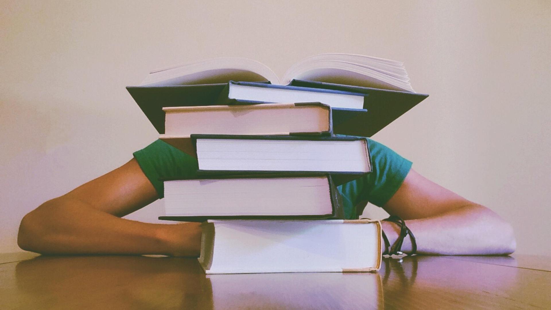 Person resting arms on a table with books stacked on their head.