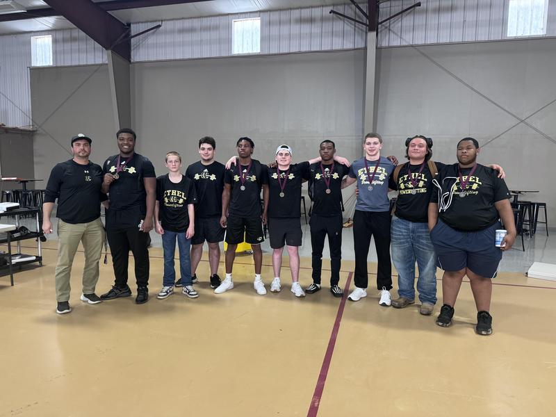 Ethel Tiger powerlifting team members and their coach stand together inside a gym facility, several athletes wearing medals around their necks after competing at the South State meet.